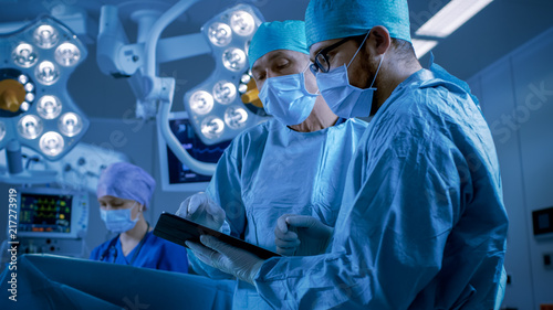 Professional Surgeons and Assistants Talk and Use Digital Tablet Computer while Standing in the Modern Hospital Operating Room.