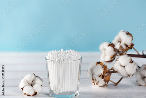 Cotton swabs in clear glass jar in bathroom setting with a cotton flowers on pastel blue background. Copy space.