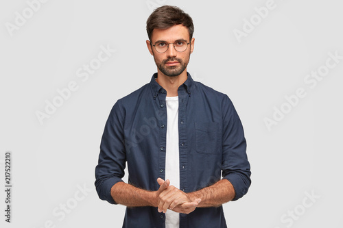 Waist up portrait of handsome serious unshaven male keeps hands together, dressed in dark blue shirt, has talk with interlocutor, stands against white background. Self confident man freelancer