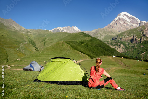 Treking for the Kazbek peak in the mountains of the Caucasus. the tourist is sitting on the grass by the tent and is looking at the snowy peak.