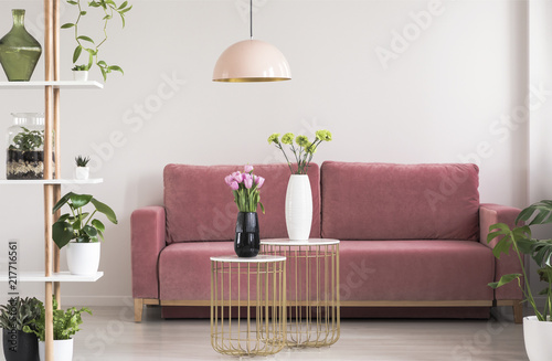Real photo of a feminine living room interior with gold and marble tables with fresh flowers standing in front of a cozy pink sofa
