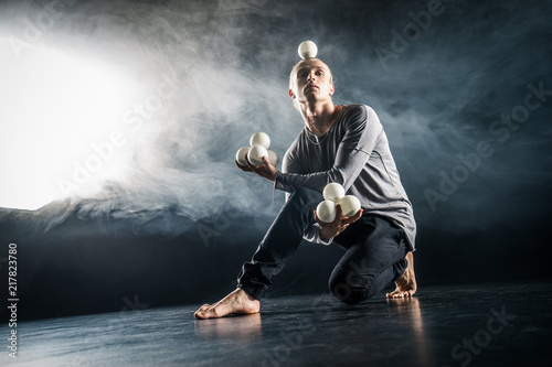 Blond juggler sitting on the floor with white balls on black background