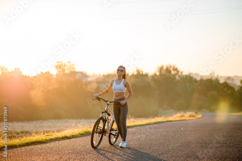 Beautiful happy fit and sporty woman in sportswear standing with bicycle on the countryside road near the field on sunset.
