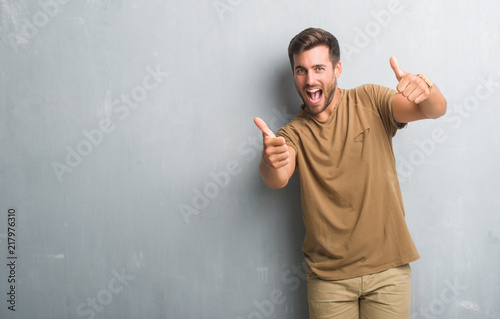 Handsome young man over grey grunge wall approving doing positive gesture with hand, thumbs up smiling and happy for success. Looking at the camera, winner gesture.