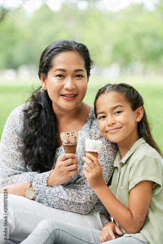Asian lady and cute girl holding tasty ice-cream and looking at camera while sitting on blurred background of park