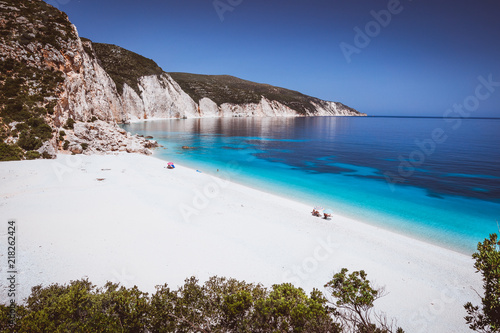 Fteri beach, Kefalonia, Greece. Lonely tourists protected from sun umbrella chill relax near clear blue emerald turquoise sea water lagoon. Framed by tree foliage