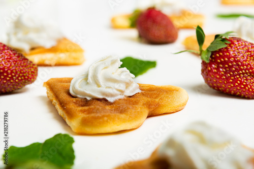 Close-up of a heart shaped waffle decorated with cream, strawberries and mint leaves on white background.
