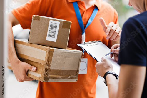 A person wearing an orange T-shirt and a name tag is delivering a parcel to a client, who is putting his signature on the receipt. Friendly worker, high quality delivery service.