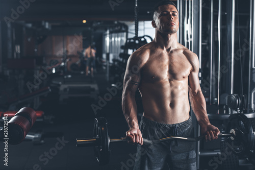 Serious shirtless sportsman is doing biceps curls while using weight. He is standing among machine zone and making efforts. Copy space in left side