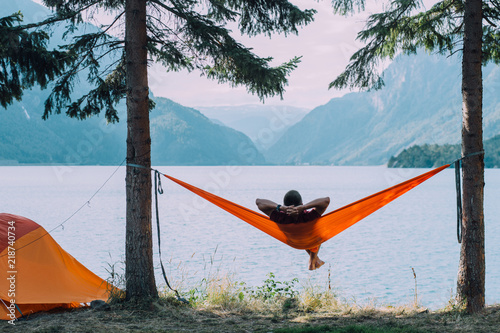 Back view of man silhouette relaxing on orange hammock between two trees pine enjoying the view at the lake in summer norwegian cloudy morning.