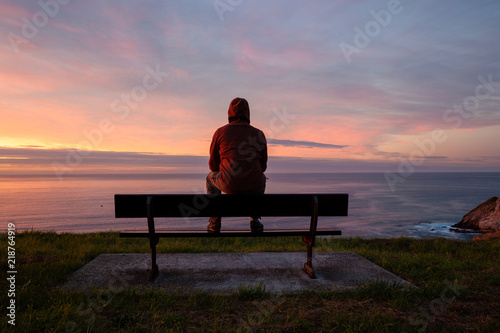 Lonely man sits alone on the rocky coast and enjoying sunset. View over rocky cliff to ocean horizon