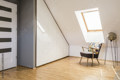 Lamp next to armchair on wooden floor in white attic interior with door and window. Real photo