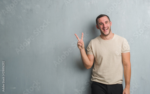 Young caucasian man over grey grunge wall smiling with happy face winking at the camera doing victory sign. Number two.