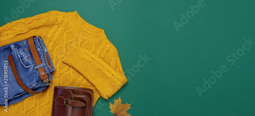 Female orange knitted sweater, blue jeans, leather bag and autumn leaves on green background top view flat lay. Fashion Lady Clothes Set Trendy Cozy Knit Jumper Autumn accessories Female fashion look
