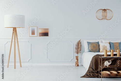 Light grey bedroom interior with gold accents in real photo with big lamp, double bed and decorations