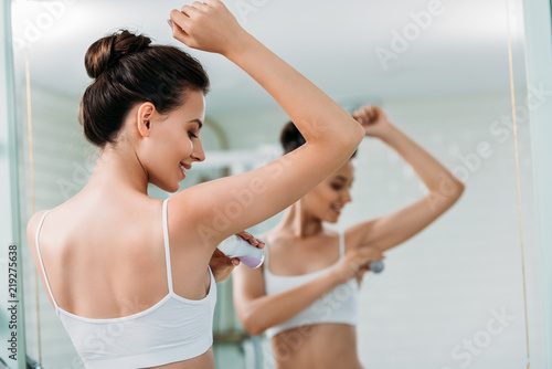 beautiful young woman applying roller deodorant at mirror in bathroom