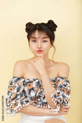 Portrait of beautiful young Asian woman with hair buns pouting lips and looking flirty at camera on light yellow background