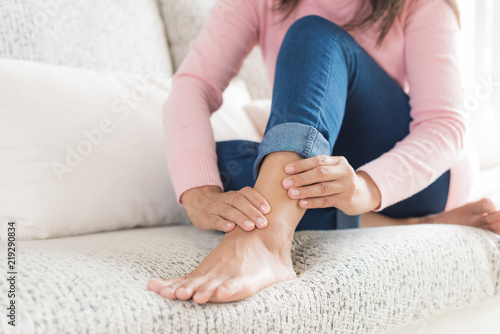 Closeup woman sitting on sofa holds her ankle injury, feeling pain. Health care and medical concept.