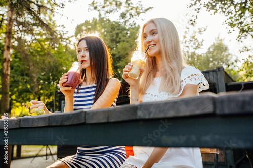 Two pensive hipster girls, women, friends the blonde and the brunette drink cocktails at modern outdoor cafe on the green park in sunset beatiful light. Wearing in striped and white dress.
