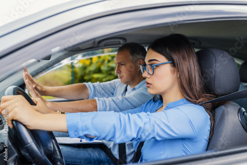  Young serious inexperienced woman driving a car in alert, worried instructor man sitting aside and looking nervous at the road, dangerous situation