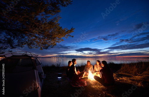 Night summer camping on sea shore. Group of five young tourists sitting on the beach around campfire near tent under beautiful blue evening sky. Tourism, friendship and beauty of nature concept.