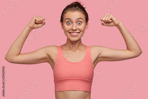 Sporty energetic athletic woman wears pink sports top smiles as shows her biceps, likes sport and active lifestyle, poses against studio background. People, health and physical activity concept