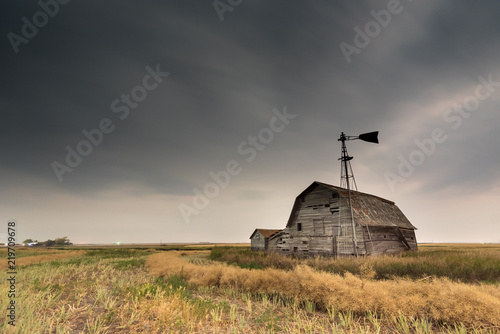 Vintage barn, bins and windmill in a swathed canola field under ominous dark skies in Saskatchewan, Canada