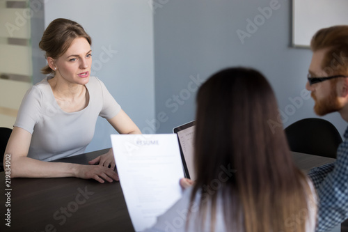 Serious millennial female job candidate talking during work interview in office, recruiters listening to applicant while hiring, hr team thinking considering employee candidature. Employment concept