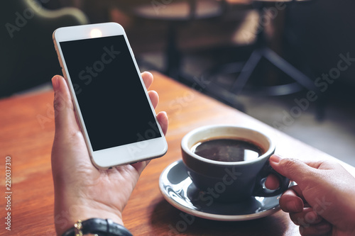 Mockup image of hands holding white mobile phone with blank black screen while drinking coffee in modern cafe