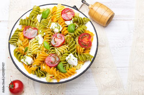 Kuchnia włoska - Sałatka z kolorowego makaronu, pomidorów koktajlowych, sera feta i świeżej bazylii na białym drewnianym tle.