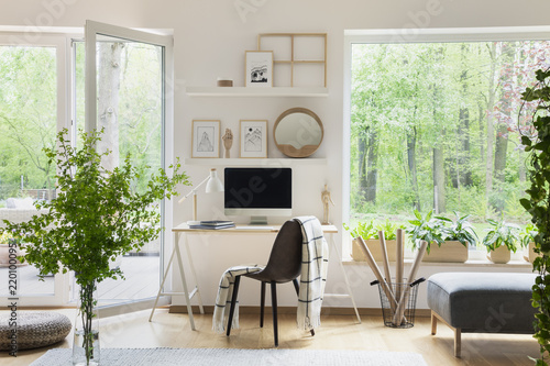 Real photo of white living room interior with big window, glass door, fresh plants, wooden desk with mockup computer and simple posters on shelves