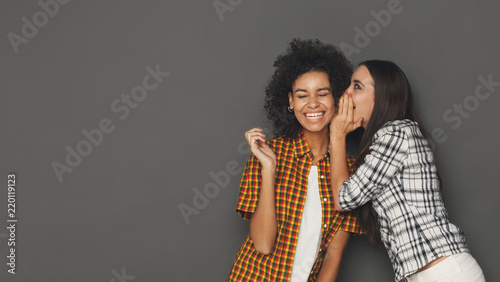 Young woman whispering to her friend against grey background