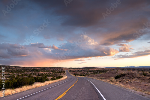 Highway curving into the distance through the landscape near Santa Fe, New Mexico underneath a dramatic colorful sky at sunset