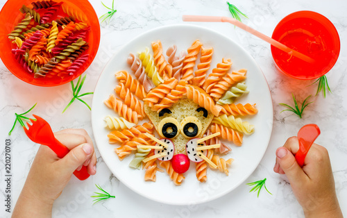 Lion pasta - fun food idea for kids lunch, animal shaped food art. Colorful fusilli vegetables pasta with sandwich like a cute lion head on white plate top view