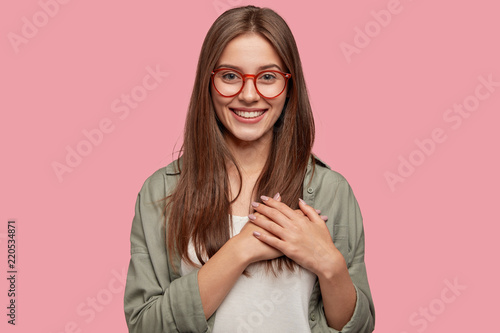 People, dedication and promise concept. Happy girlfriend keeps hands on heart, swears be loyal to boyfriend, expresses gratitude, wears spectacles, casual shirt, stands against pink studio wall.