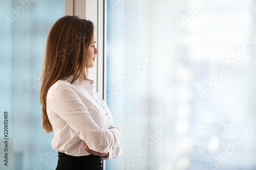 Successful thoughtful woman business leader looking out of big window enjoying city view and wellbeing feeling motivated for new goals, rich millennial businesswoman dreaming of future, copy space