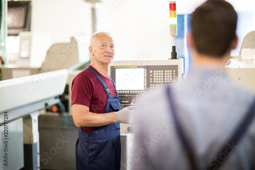 Senior engineer consulting his young subordinate while standing by industrial machine in workshop