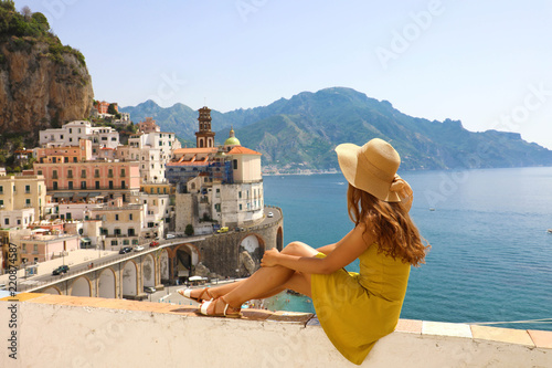 Beautiful young woman with hat sitting on wall looking at stunning panoramic village of Atrani on Amalfi Coast, Italy