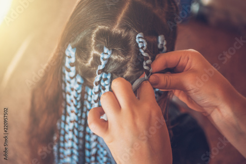 Hairdresser weaves braids with kanekalon material to young girl head, making creative hairstyle with thick plaits or pigtails also known as Afro braids