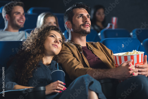 young couple with popcorn holding hands while watching movie together in cinema