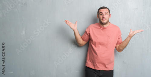 Young caucasian man over grey grunge wall clueless and confused expression with arms and hands raised. Doubt concept.