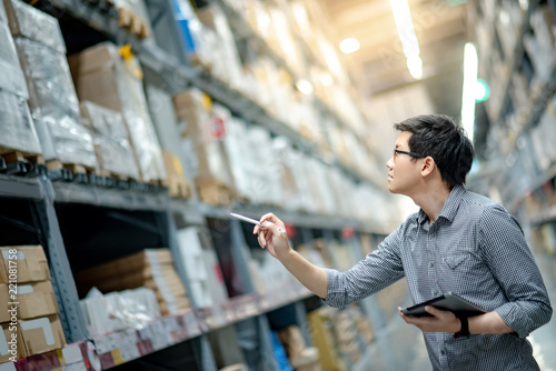 Young Asian man worker doing stocktaking of product in cardboard box on shelves in warehouse by using digital tablet and pen. Physical inventory count concept