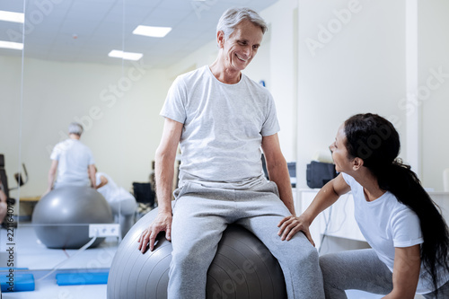 Funny ball. Positive enthusiastic aged man feeling excited while spending time in a professional rehabilitation center and sitting on a comfortable big fitball
