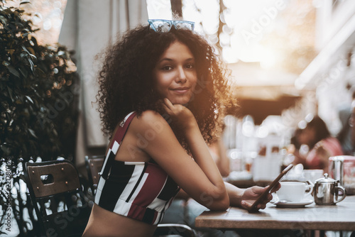 Cute curly African-American female in a crowded street bar holding a smartphone, with the cup of a hot tea and a metal teapot in front; young Moroccan girl in street cafe with the cellphone