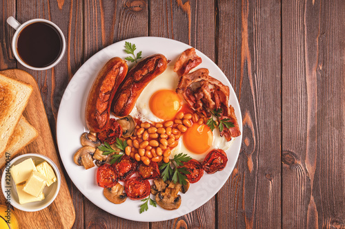 Traditional full English breakfast with fried eggs, sausages, beans, mushrooms, grilled tomatoes and bacon on wooden background.