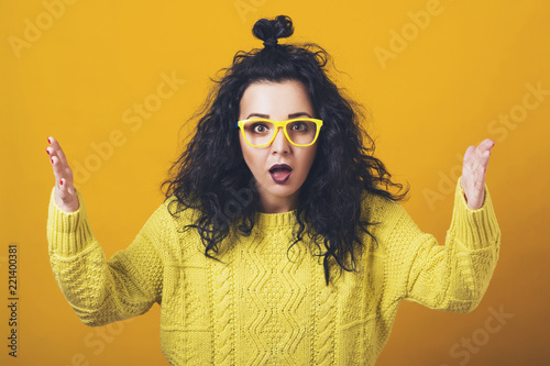Shocked amazed young woman, standing yellow background. Human emotions, facial expression concept. Girl with curly hair in glasses, yellow knitted sweater. Horrible, stress, shocked.