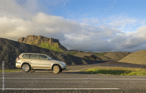 dirty off road car standing next asphalt road with Iceland northern summer landscape, green eroded mountains and blue sky, white clouds