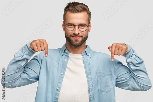 Lets go downstairs. Photo of attractive unshaven man with appealing look, points down with both index fingers, dressed in casual shirt, isolated over white background. Advertisement concept.