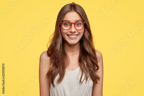 Indoor shot of good looking young Caucasian woman has broad smile, being in good mood after date with boyfriend, wears round glasses, looks directly at camera, poses against yellow studio wall.