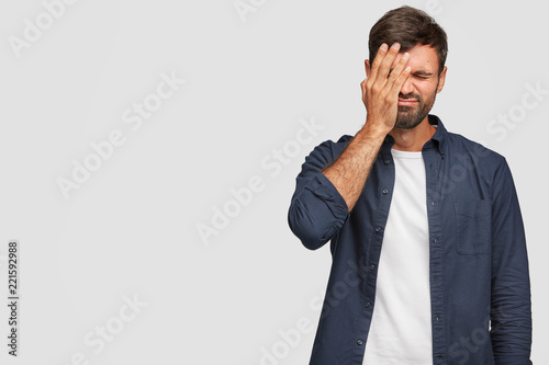 Indoor shot of handsome stressful overworked man covers face with palm, has displeased expression, dressed in casual clothes, poses against white background with copy space for your promotional text